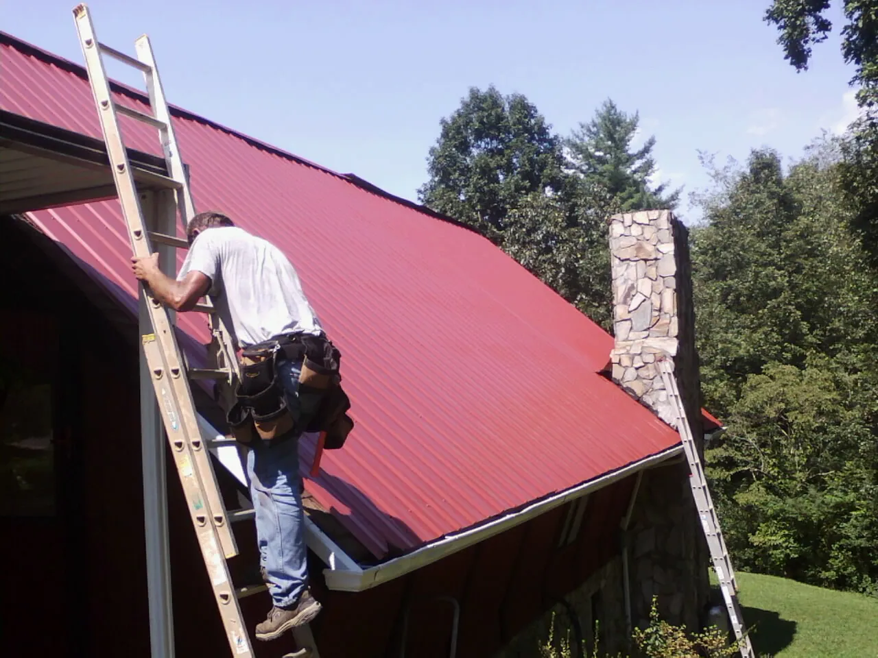 Roofer on a ladder beside a red metal roof and stone chimney — Soffit & Fascia Repair by Roofing Brothers in Sioux Center